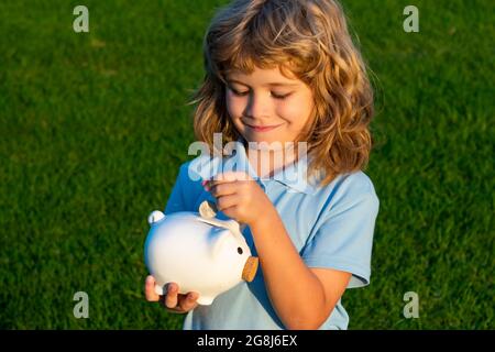 Child boy putting in piggy bank coin money. Portrait of little boy with moneybox piggybank on grass background. Stock Photo