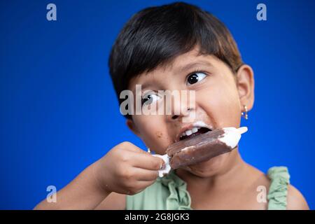 Cloe up head shot of girl kid busy eating ice cream on blue studio background - concept of unhealthy food consumption Stock Photo