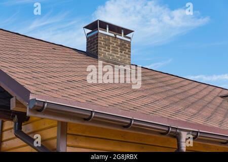 Bitumen asphalt roofing shingles and brick chimney pipe on a wooden house. Individual heating system. Rain gutter. Stock Photo