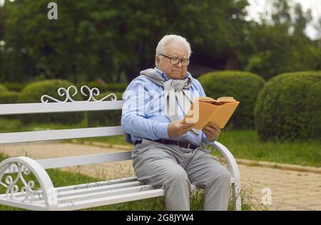 Elderly senior man in eyeglasses enjoy reading book sitting on bench in park Stock Photo