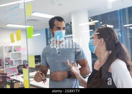 Two diverse creative colleagues wearing face masks talking at meeting Stock Photo