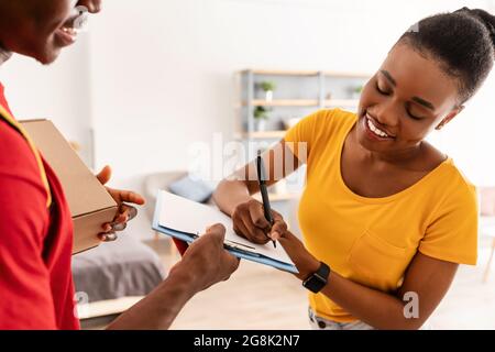 Black Lady Signing Papers Receiving Parcel Box From Courier Indoors Stock Photo