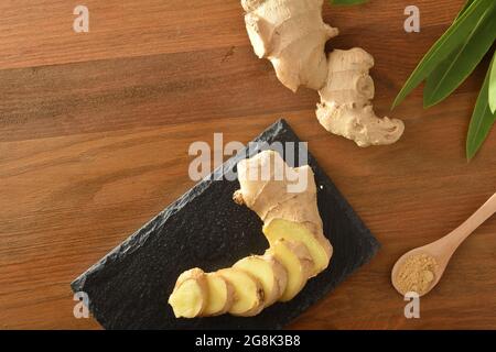 Ginger root slice on slate plate and spoon with powder on wooden table with root and leaves. Top view. Stock Photo