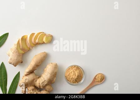 Ginger root and with slices and powder in glass jar and wooden spoon on white table with leaves. Top view. Stock Photo