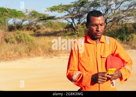 African male mechanic checking his cell phone with helmet on his arm. Concept of engineer or technician. Stock Photo