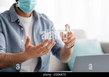 Unrecognizable man in face mask disinfecting hands with disinfectant mist spray while using modern laptop Stock Photo