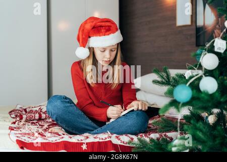 A teenager girl in a Christmas cap sitting on a bed at home writes a letter to Saint Claus Stock Photo