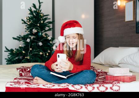 A red-haired teenager girl in a Santa hat talking on a video chat on the mobile phone. Stock Photo