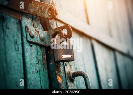 An old rusty lock hangs on the abandoned wooden gate, painted blue, which closes them and is illuminated by sunlight. Secret. Stock Photo