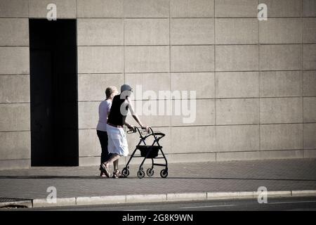 Old couple walking in a street near a grey wall. Old man with walker. Stock Photo