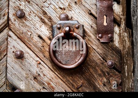 Heavy rusty iron door knocker, ring in front of rectangular iron plate on weathered grained massive old wooden door at the entrance of castle complex Stock Photo