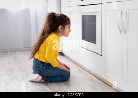 Cute little girl sitting near oven in kitchen and looking inside Stock Photo