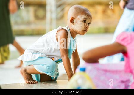 Young Burmese child with thanaka on face and shaved head climbs on wall, Shwedagon Pagoda, Yangon, Myanmar Stock Photo