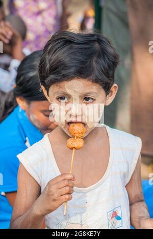 Cute young Burmese child with thanaka yellow paste face powder eating meat on stick, Bagan, Myanmar Stock Photo