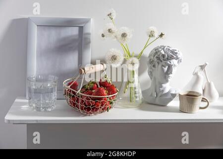 A bunch of fluffy dandelions in a chemical flask, an empty photo frame, a cup of espresso coffee, and a plaster head of David on a white chest of draw Stock Photo