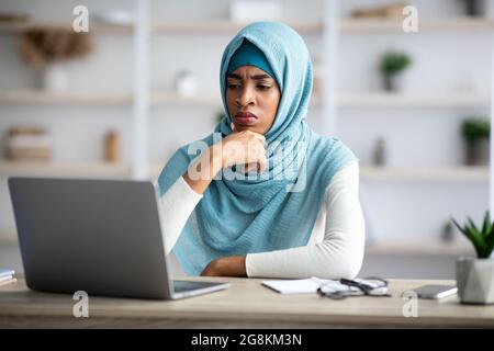 Stressed black muslim freelancer lady looking at laptop screen with worry Stock Photo