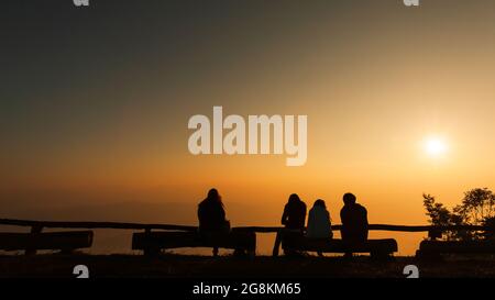 A group of tourists sits on the logs at viewpoint while  relaxing on a mountain peak at sunrise, the rising sun over a mountains in the backgrounds. Stock Photo
