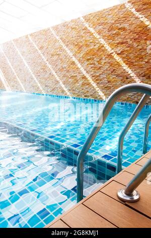 An empty turquoise swimming pool with grab bars ladder at sunrise. Focus on grab bars ladder. Stock Photo