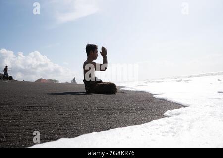 A Balinese Hindu man praying on the beach on a hot sunny day. The sand is black and the waves are foamy white Stock Photo