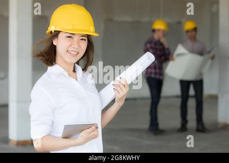 Female Asian enginee holding blueprint plan and tablet computer in hands standing and smiling with selt confident while two male team talking in blur Stock Photo
