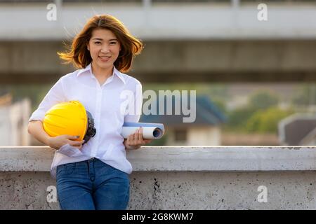 Cute smiling face of Asian female engineer holding blueprint and yellow safty helmet looking to camera with self confident manner on rooftop of constr Stock Photo