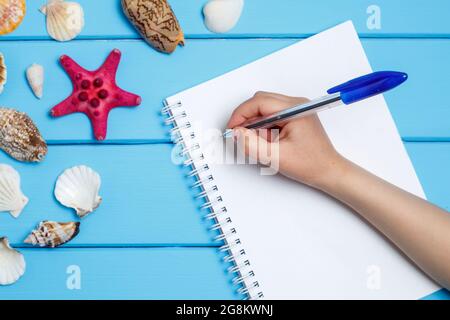 Blank notepad and hand with a pen different marine items on blue painted wooden background. Sea objects on wooden planks. Selective focus. Place for t Stock Photo