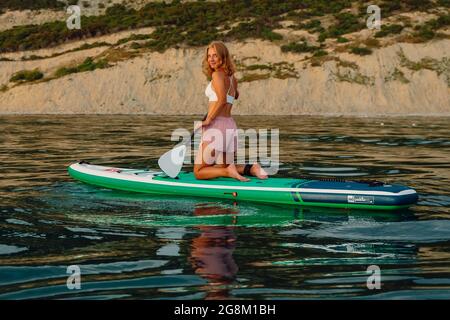 June 25, 2021. Anapa, Russia. Slim girls on stand up paddle board at quiet sea. Women sit on Red Paddle SUP board in sea. Stock Photo