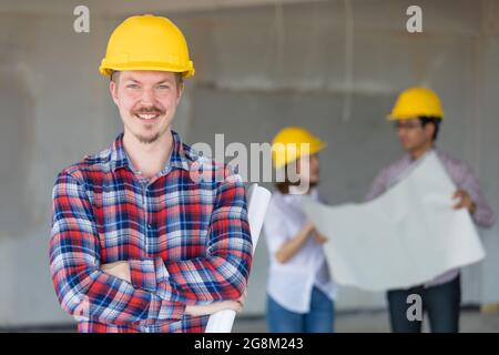 Caucasian engineer wearing yellow safty hat standing and looking to camera. There are two engineers talking in blur background. Stock Photo