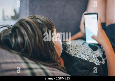 Teen boy checking social media holding smartphone, using mobile phone app playing game, relaxing on sofa at home. Emotional isolation and depression Stock Photo