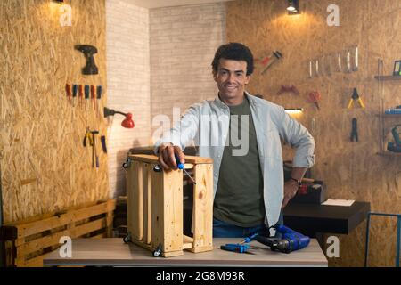 Portrait of a smiling carpenter with screwdriver in workshop with tools and accessories in the background. Stock Photo