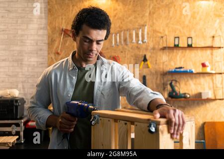 Carpenter works with a drill on a small wooden crate in workshop. Stock Photo