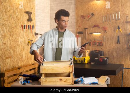 Carpenter in workshop with one hand measuring the wooden box , while holding and looking at the tablet in the other hand. Stock Photo