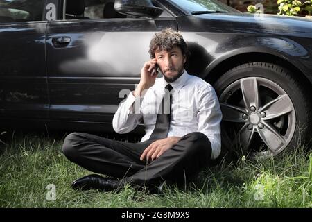 Portrait of a tired Italian businessman sitting on the ground against a car and talking on the phone Stock Photo