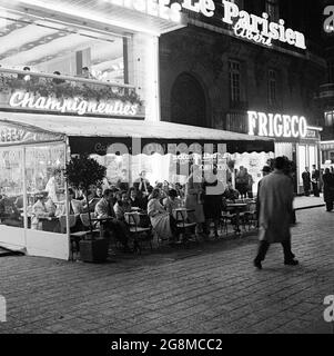 1950s, historical, cafe society....evening-time in Paris and parisians enjoy a drink and conversation in an open-air covered area outside a cafe on the sidewalk of the Avenue des Champs-Elysées. A retail store selling 'Frigeco' refrigerators can be seen in the picture. Stock Photo
