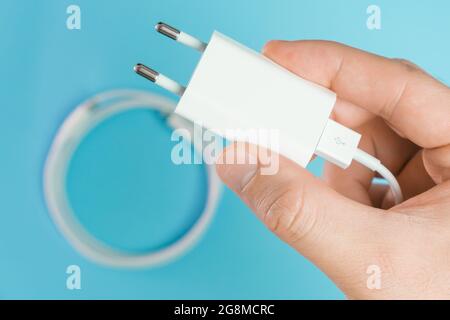 A white smart phone charger cable with lightning hi speed USB type A connector and adapter in man hand on the blue background. Stock Photo