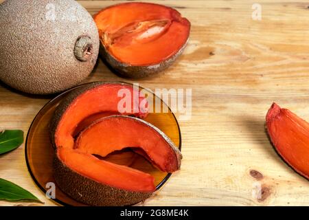 Slices of ripe sapodilla plum on a brown crystal dish on a wooden table, there is also a whole fruit nearby, and a half cut next to it Stock Photo
