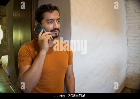 Handsome bearded Hispanic man with an orange t-shirt leaning on the wall and talking on the phone Stock Photo