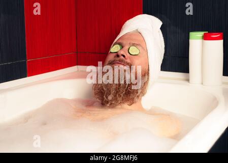 Cute bearded man taking bath with head wrapped in towel and cucumber slices on his eyes. Funny hipster relaxing in foamy bathtub and enjoying life Stock Photo