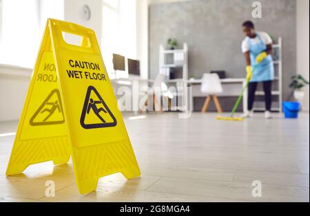 Close up of sign warning about wet floor placed by janitor who's working in background Stock Photo