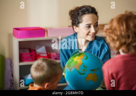 back view of blurred boys near globe and smiling teacher in montessori school Stock Photo