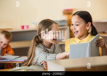 excited girl pointing at book near cheerful asian friend in montessori school Stock Photo