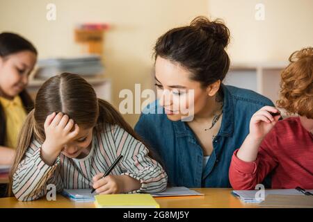 young teacher looking at girl writing in notebook during lesson Stock Photo