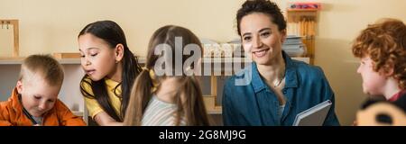 happy young teacher smiling near interracial kids in montessori school, banner Stock Photo