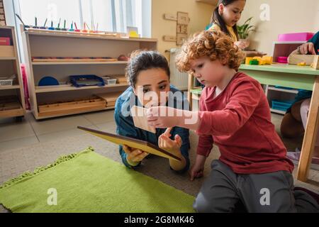 teacher and boy playing with wooden boards on floor in montessori school Stock Photo