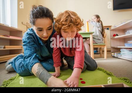 tattooed teacher playing on floor with redhead boy near girl on blurred background Stock Photo