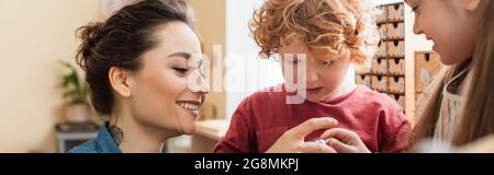 happy teacher near curly boy and blurred girl in montessori school, banner Stock Photo