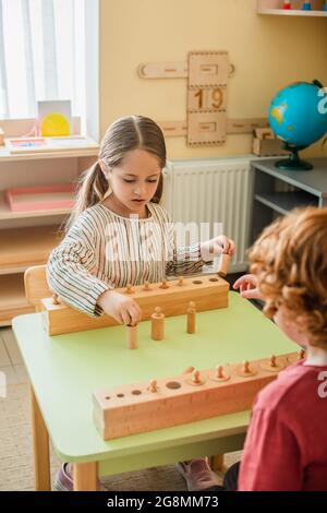 girl playing game with wooden element near blurred kid in montessori school Stock Photo