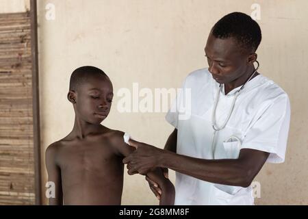 Doctor helping young African child to heal from vaccine shot in the upper shoulder. Wearing lab coat and having a stethoscope. Stock Photo