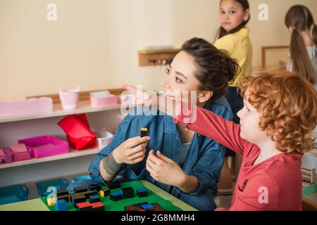redhead boy pointing with finger near smiling teacher holding wooden cube Stock Photo