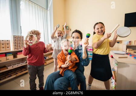 cheerful interracial kids with young teacher playing musical instruments in montessori school Stock Photo